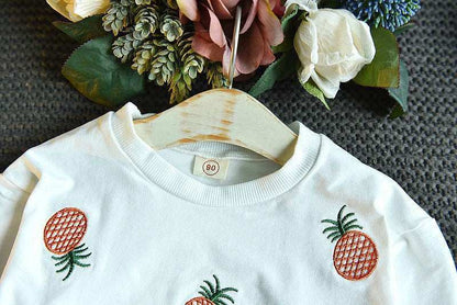 Embroidered sweater with pineapple design paired with a floral backdrop and hanger.